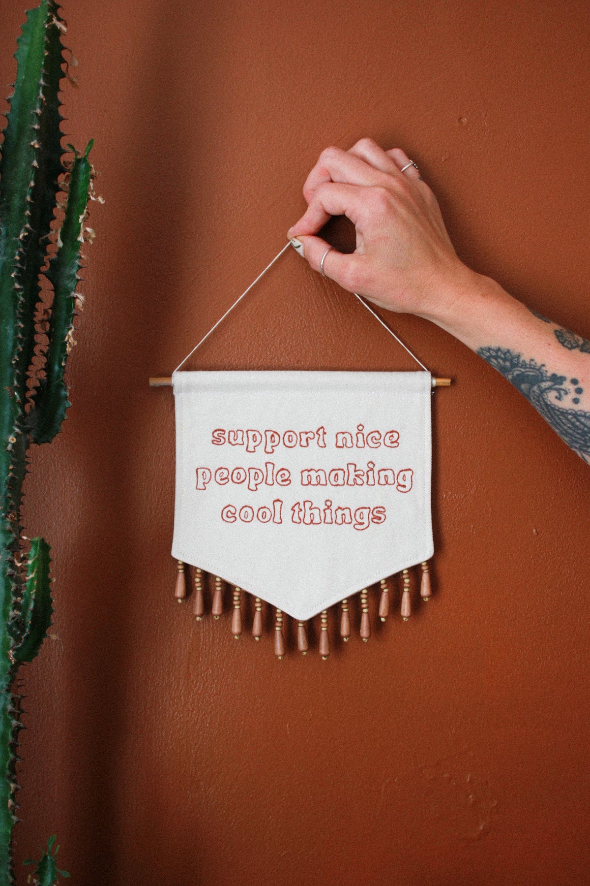 A hand holds a small white fabric wall banner with wooden dowels and fringe, embroidered with the phrase 'support nice people making cool things' in red thread, against a terracotta wall with a cactus on the left.