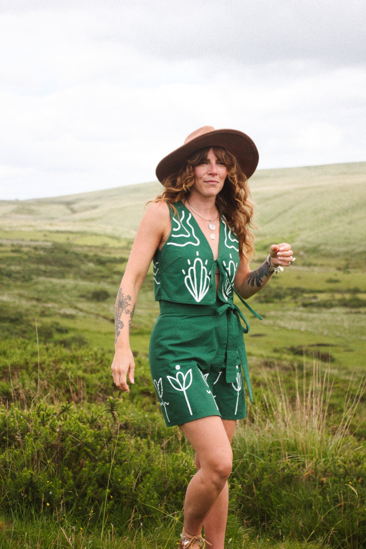 A woman wearing a green sleeveless romper with white botanical line drawings and a brown wide-brim hat, standing in a grassy outdoor landscape.