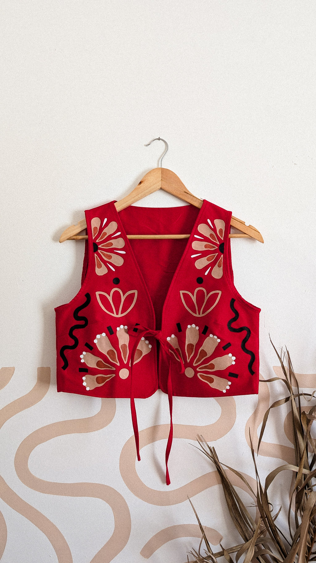 A red sleeveless vest with beige and black floral embroidery, hanging on a wooden hanger against a white wall with a beige wavy pattern and dried grass in the foreground.