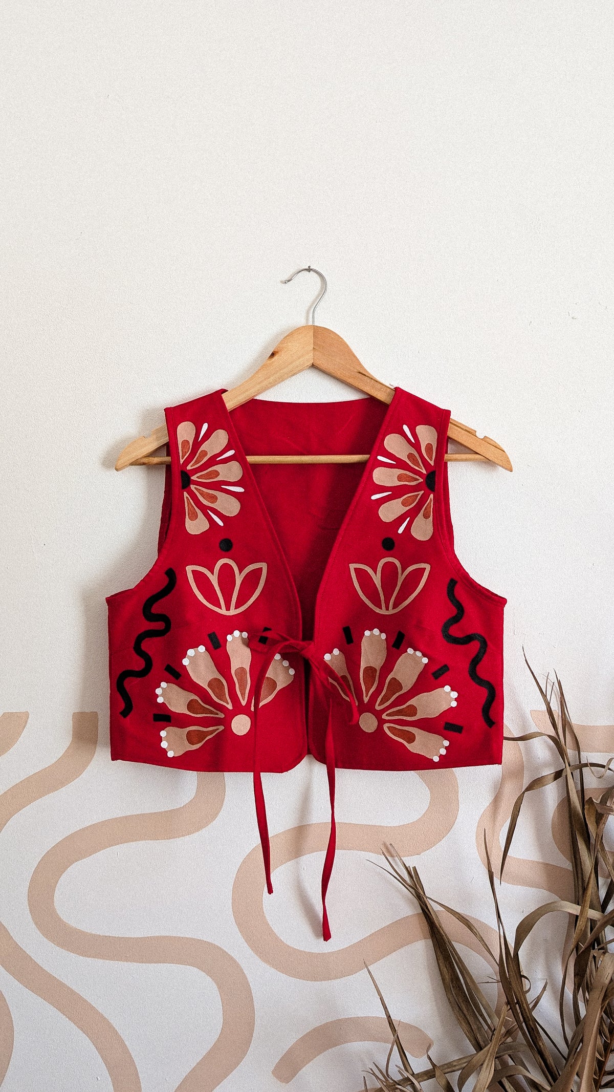 A red sleeveless vest with beige and black floral embroidery, hanging on a wooden hanger against a white wall with a beige wavy pattern and dried grass in the foreground.